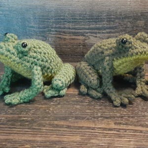 May include: Two handmade crochet frogs in shades of green and yellow sit side-by-side. The frogs have detailed features, including bulging eyes and textured skin. They are posed on a wooden surface, showcasing their lifelike appearance.