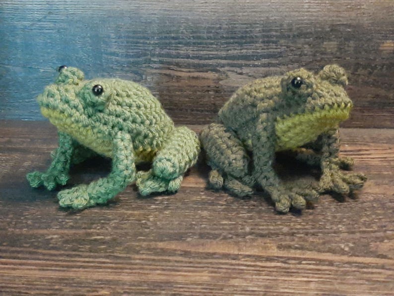 May include: Two handmade crochet frogs in shades of green and yellow sit side-by-side. The frogs have detailed features, including bulging eyes and textured skin. They are posed on a wooden surface, showcasing their lifelike appearance.