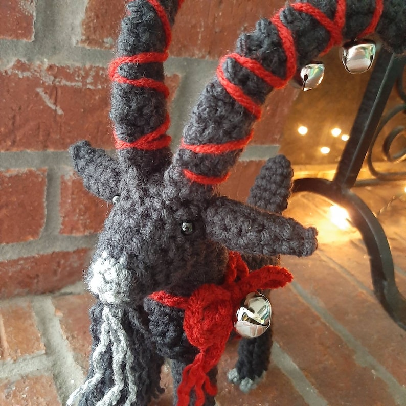 May include: A gray crocheted goat with red yarn accents and silver bells. The goat has a long, white beard and a red scarf around its neck.