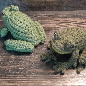 May include: Two handmade crochet frogs in shades of green. One frog is upright with its legs extended, and the other is in a sitting position. Both frogs have black bead eyes and are displayed on a wooden surface.