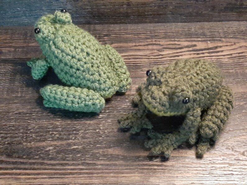 May include: Two handmade crochet frogs in shades of green. One frog is upright with its legs extended, and the other is in a sitting position. Both frogs have black bead eyes and are displayed on a wooden surface.