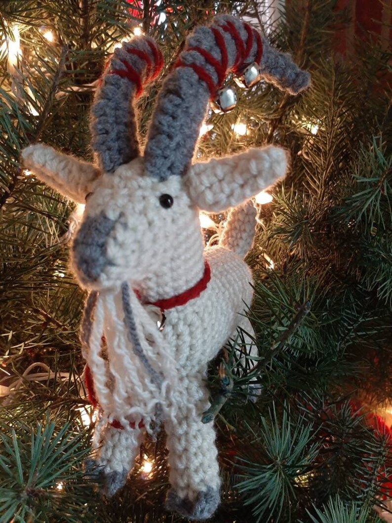May include: A white crocheted goat ornament with gray horns and a red and white striped scarf. The goat has a white beard and is wearing jingle bells on its horns.