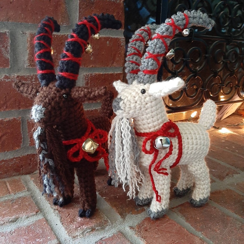 May include: Two crocheted goat ornaments with red and white ribbon bows and bells. The goat on the left is brown with black horns and the goat on the right is white with gray horns.