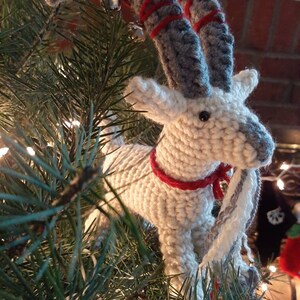 May include: A white crocheted goat ornament with gray horns and red stripes. The goat is wearing a red ribbon around its neck and has a gray and white scarf. The ornament is hanging on a green Christmas tree.