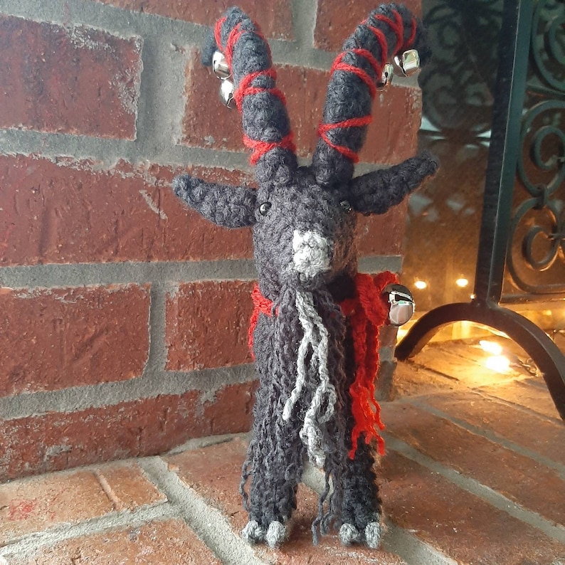 May include: A gray crocheted goat with red and white yarn horns and a red scarf. The goat has a white beard and is standing in front of a brick wall.