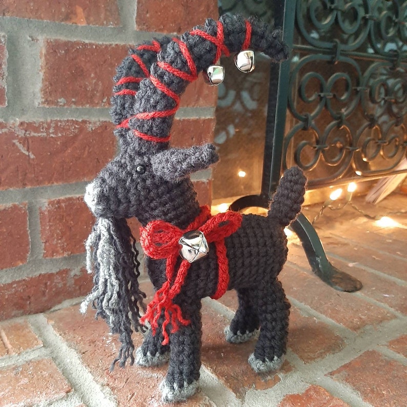 May include: A crocheted black goat with red and white ribbon and bells. The goat has a long, gray beard and is standing on a brick surface.