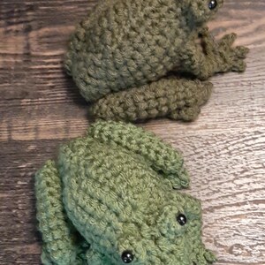May include: Two handmade crochet frogs in shades of green. The frogs have detailed features, including bulging eyes and textured skin. The frogs are positioned on a wooden surface, showcasing the craftsmanship of the crochet work.