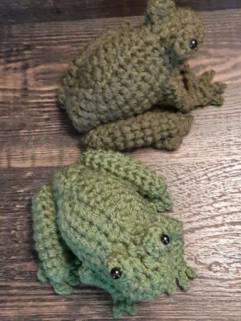 May include: Two handmade crochet frogs in shades of green. The frogs have detailed features, including bulging eyes and textured skin. The frogs are positioned on a wooden surface, showcasing the craftsmanship of the crochet work.