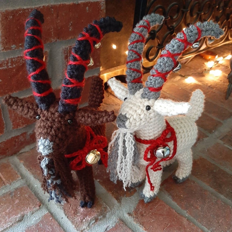 May include: Two crocheted goat ornaments with red and white ribbon and bells. The goat on the left is brown with black and red yarn. The goat on the right is white with gray and red yarn.