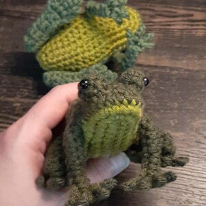 May include: Two handmade crochet frogs in shades of green and yellow. One frog is held in a hand, showcasing its detailed features, including black bead eyes and textured limbs. The other frog is in the background, also crocheted.