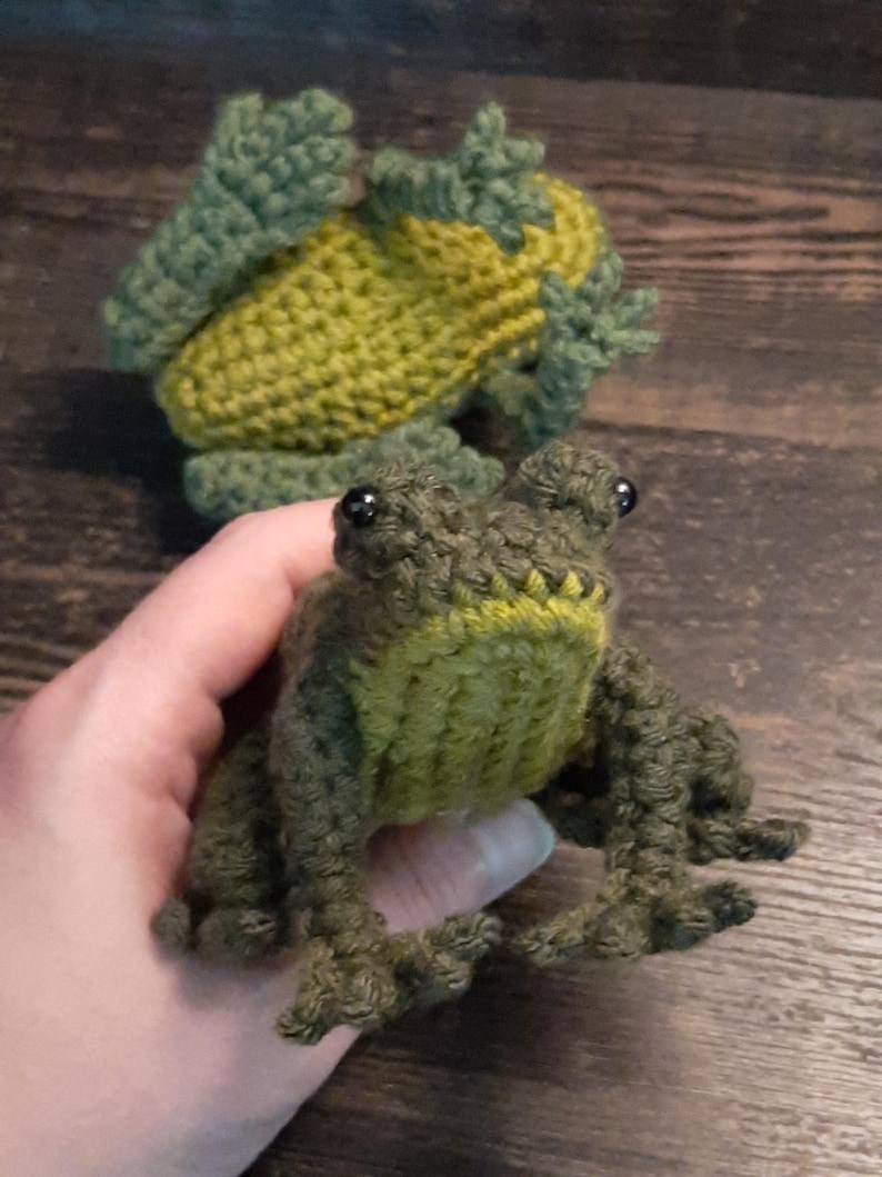 May include: Two handmade crochet frogs in shades of green and yellow. One frog is held in a hand, showcasing its detailed features, including black bead eyes and textured limbs. The other frog is in the background, also crocheted.
