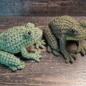 May include: Two crocheted frog figurines. One is light green, the other is olive green. Both have black eyes and detailed legs and feet. The frogs are sitting on a wooden surface.