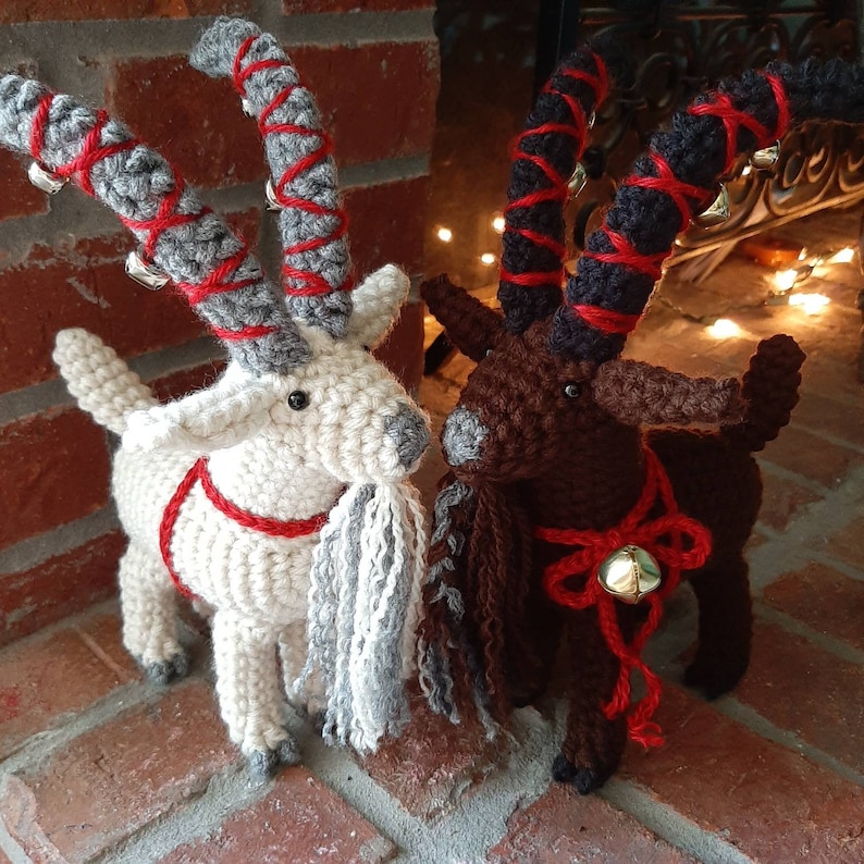 May include: Two crocheted goat ornaments with red and white accents. The goats have long, curly horns and are wearing red and white ribbon collars with bells.