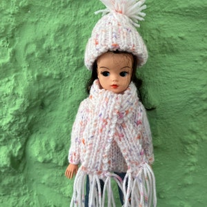 May include: A white knitted doll sweater with a matching white knitted hat and scarf with a pom-pom on top. The sweater and scarf have a speckled pattern with pink, orange, and purple accents.