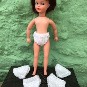 May include: A white doll wearing a white knitted nappy. Four additional white knitted nappies are laid out on a black surface.