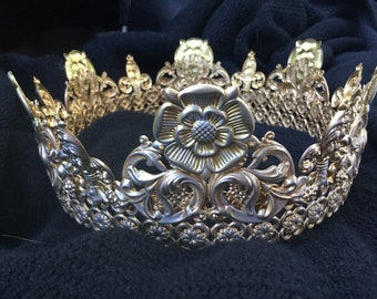 Puede incluir: Una corona dorada con un gran diseño floral ornamentado en el centro. La corona está hecha de metal y tiene un diseño delicado e intrincado.
