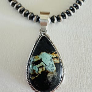 May include: A teardrop-shaped pendant featuring a black stone with light blue and brown matrix veins, set in a silver frame with twisted wire detailing, suspended from a necklace of round, polished silver beads with dark oxidized accents.
