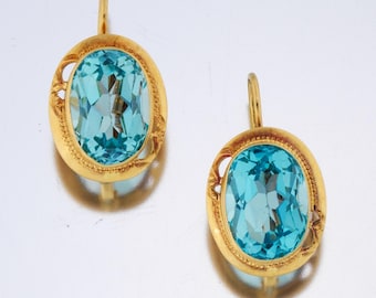 May include: A pair of gold-toned earrings featuring large, oval, light blue gemstones. The gemstones are set in ornate gold frames with decorative cutouts. The earrings have a classic, elegant design.