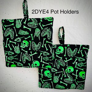 May include: Two black pot holders with a repeating pattern of glowing green skeletons, skulls, and bones. Each pot holder has a small loop for hanging and the text "2DYE4 Pot Holders" is printed on the top.