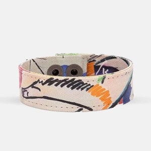 Op de afbeelding: Een kleurrijk stoffen armband met een witte achtergrond en een abstract ontwerp in zwart en oranje. De armband heeft twee metalen gespen.