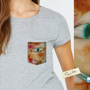 May include: A grey t-shirt with a pocket featuring a painting of a cat's face with a green eye. The text "Paul Klee" is written on a white sticker on the shirt.