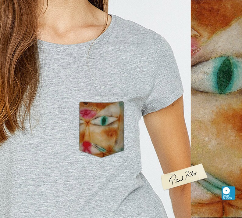May include: A grey t-shirt with a pocket featuring a painting of a cat's face with a green eye. The text "Paul Klee" is written on a white sticker on the shirt.