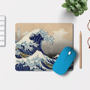 Può includere: Un tappetino per mouse rettangolare con l'iconica opera d'arte giapponese "La grande onda di Kanagawa" nei toni del blu e beige. Un mouse wireless blu è sul tappetino. Sono visibili anche una tastiera, una matita, occhiali e una piccola pianta.
