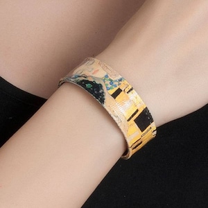 May include: A wide cuff bracelet featuring a print of Gustav Klimt's "The Kiss." The bracelet has a gold, black, and yellow colour scheme, with a portrait of a couple embracing. The bracelet is worn on the wrist.
