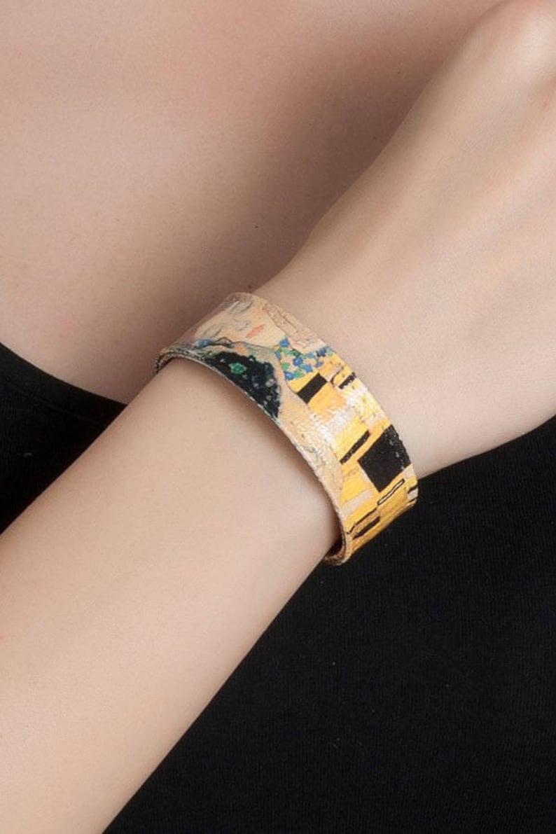 May include: A wide cuff bracelet featuring a print of Gustav Klimt's "The Kiss." The bracelet has a gold, black, and yellow colour scheme, with a portrait of a couple embracing. The bracelet is worn on the wrist.
