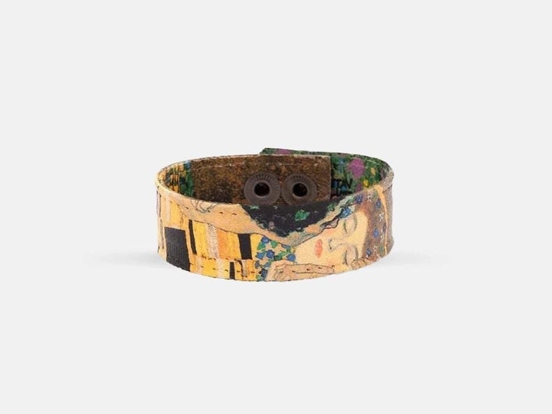 May include: A wide cuff bracelet featuring a print of Gustav Klimt's "The Kiss." The bracelet has a brown leather backing and two dark metal snaps. The artwork includes gold, yellow, black, and blue hues.