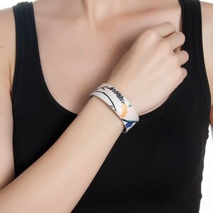 Op de afbeelding: Een witte leren armband met een kleurrijk abstract ontwerp. De armband is breed en heeft een eenvoudige, moderne stijl.