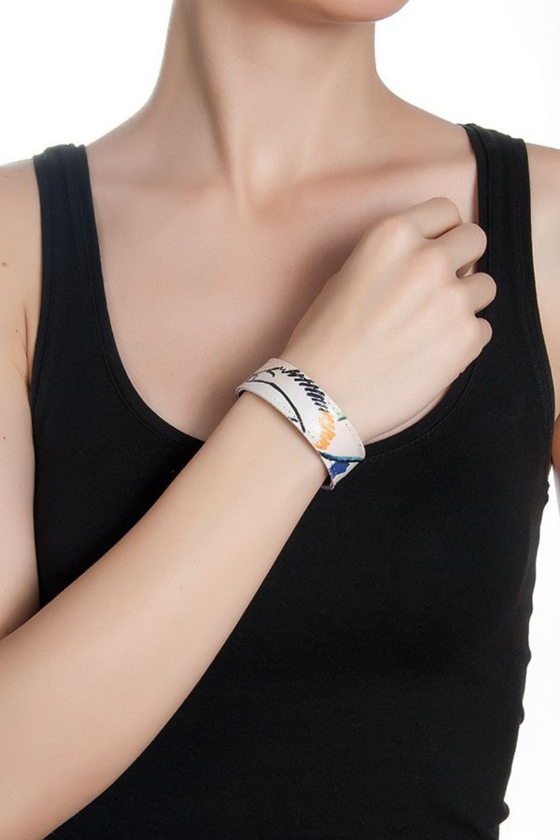 Op de afbeelding: Een witte leren armband met een kleurrijk abstract ontwerp. De armband is breed en heeft een eenvoudige, moderne stijl.