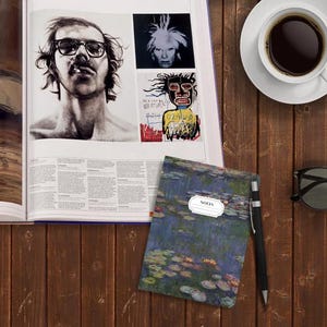 May include: A blue notebook with a water lily print and the word "NOTES" in a white circle on the cover. The notebook is on a wooden table with a cup of coffee, a book, and sunglasses.