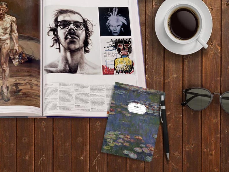 May include: A blue notebook with a water lily print and the word "NOTES" in a white circle on the cover. The notebook is on a wooden table with a cup of coffee, a book, and sunglasses.