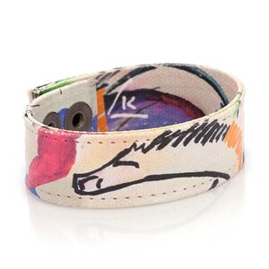 Op de afbeelding: Een kleurrijke canvas armband met een witte achtergrond en een zwart-witte tekening van een paard. De armband heeft twee zilveren drukknopen en is gestikt met wit garen.