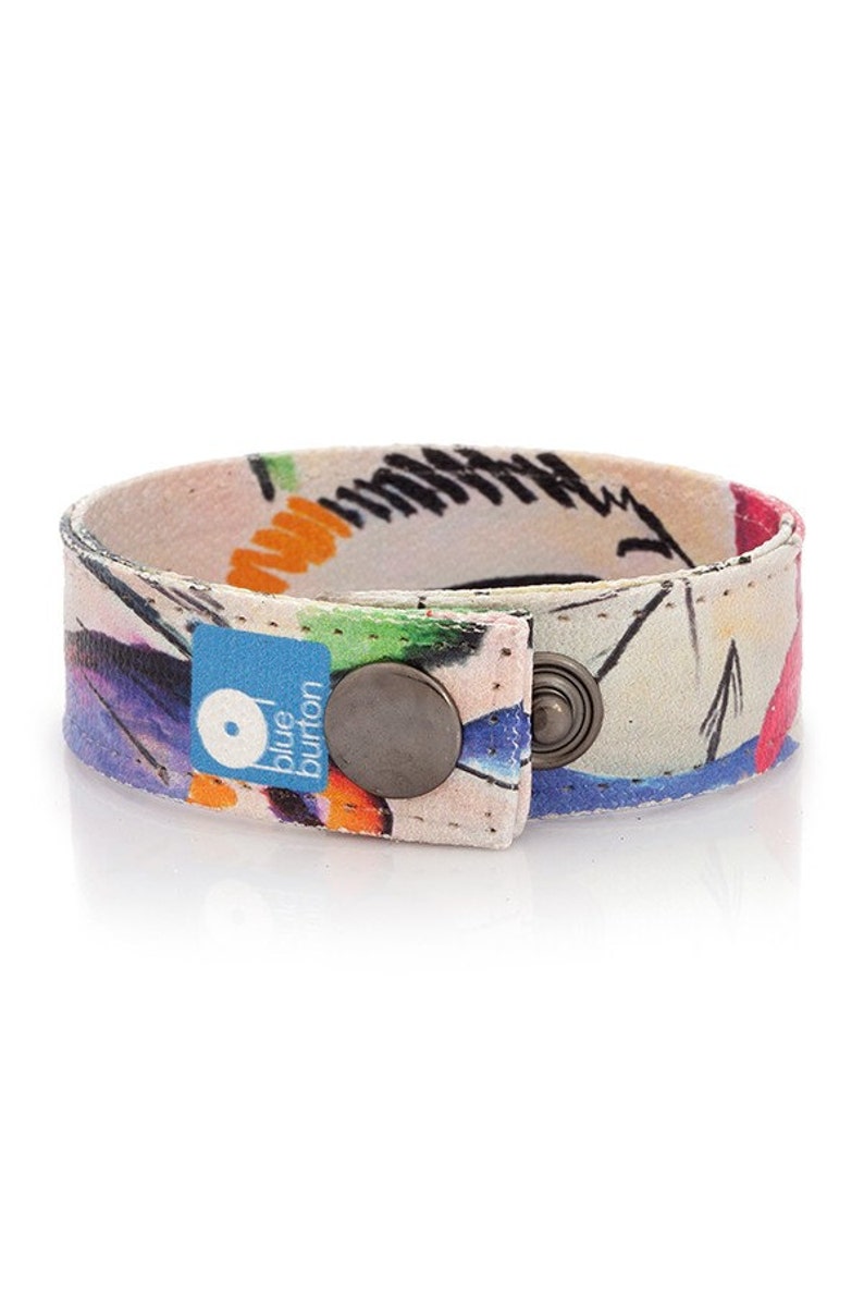 Op de afbeelding: Een kleurrijk stoffen armband met een witte achtergrond en een blauwe knoopsluiting. De knop heeft de tekst "blue button" erop.