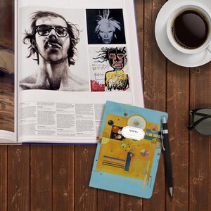 May include: A blue notebook with a yellow, blue, and black abstract painting on the cover. The notebook has a white label with the word "NOTES" printed on it. The notebook is lying on a wooden surface with a cup of coffee, a pair of sunglasses, and a pen.