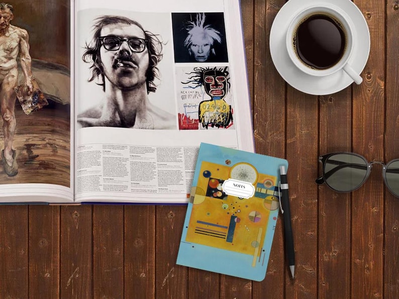 May include: A blue notebook with a yellow, blue, and black abstract painting on the cover. The notebook has a white label with the word "NOTES" printed on it. The notebook is lying on a wooden surface with a cup of coffee, a pair of sunglasses, and a pen.