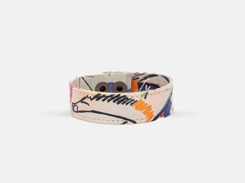 Op de afbeelding: Een kleurrijk stoffen armband met een witte achtergrond en een abstract ontwerp in zwart en oranje. De armband heeft twee metalen gespen.