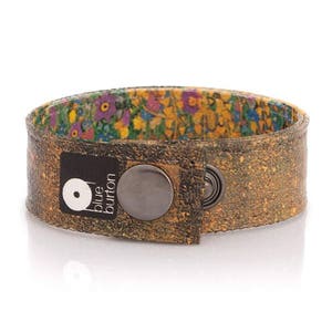 May include: A brown cork bracelet with a floral interior lining. The bracelet has a large, dark metal snap closure and a black label with the text "blue burton" and a logo. The bracelet is approximately 3.8 cm wide.