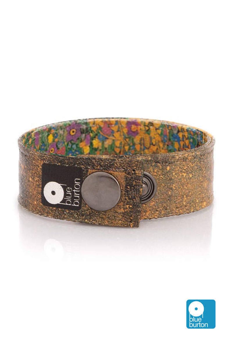 May include: A brown cork bracelet with a floral interior lining. The bracelet has a large, dark metal snap closure and a black label with the text "blue burton" and a logo. The bracelet is approximately 3.8 cm wide.