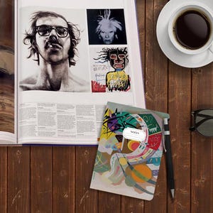 Pu&ograve; includere: Un piccolo quaderno colorato con arte astratto sulla copertina. Il quaderno ha un'etichetta bianca con la parola "NOTES" stampata su di essa. Il quaderno &egrave; appoggiato su una superficie di legno accanto a una tazza di caff&egrave;, un libro e un paio di occhiali da sole.