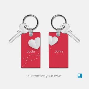 May include: Two red rectangular keychains with white paper hearts and names "Jude" and "John" printed on them. The keychains are attached to silver key rings with keys.