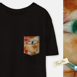 May include: Black t-shirt with a pocket featuring a colourful abstract cat painting by Paul Klee. The pocket is on the left side of the shirt. The painting is in a style reminiscent of cubism.