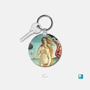 May include: A round keychain featuring a reproduction of Sandro Botticelli's "The Birth of Venus" painting. The painting depicts a nude woman standing on a seashell, with her hair flowing in the wind. She is holding a red drapery in her left hand. The background is a blue sky with clouds.