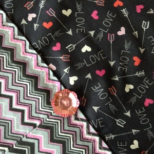 May include: A black fabric with a white chevron pattern is sewn to a black fabric with a repeating pattern of white arrows, hearts, and the word "LOVE". A pink button is sewn to the fabric.