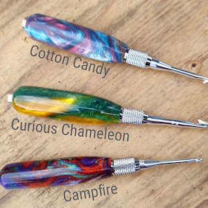 May include: Three crochet hooks with colorful resin handles. The handles are labeled "Cotton Candy", "Curious Chameleon", and "Campfire".