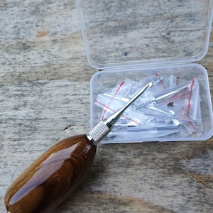 Op de afbeelding: Een haaknaald met een gepolijste houten handgreep en een zilveren metalen punt. Een doorzichtige plastic container bevat nog een aantal haaknaalden in kleine plastic zakjes. De houten handgreep heeft een glanzende afwerking.