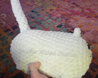 May include: White crocheted plush toy shaped like a cat with a long tail. The toy is sitting on a colorful rug. The text "Another Wicked Creation" is visible on the toy.