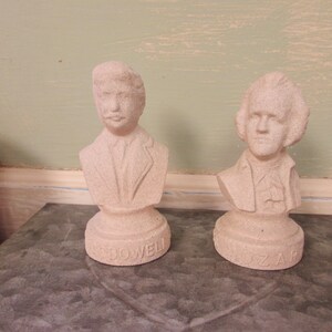 May include: Two small, beige plaster busts of famous composers. The bust on the left is labeled "Dowel" and the bust on the right is labeled "Mozart".